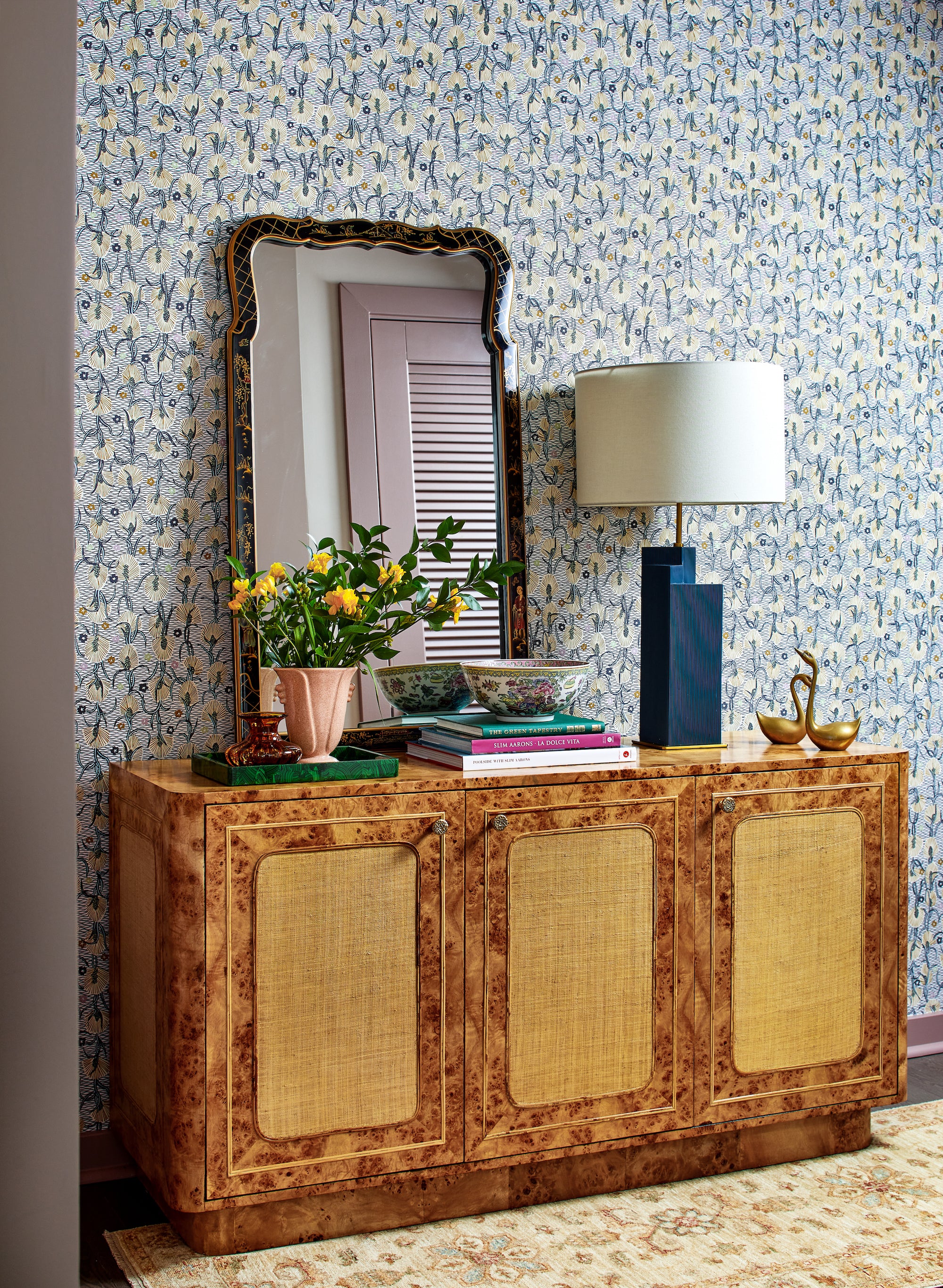 An entryway vignette featuring a burlwood console against patterned wallpaper, by Lisa & Leroy.